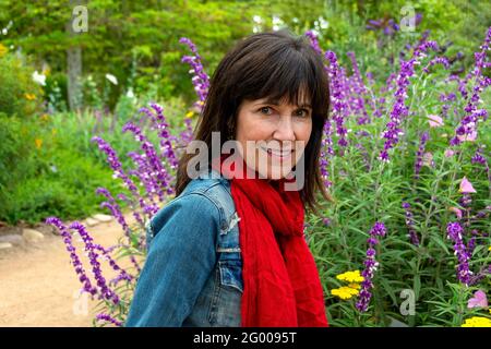 A beautiful, modern, smiling senior woman with thick brunette hair, dressed in a bright red scarf and jeans jacket, is surrounded by lush flowers in a Stock Photo