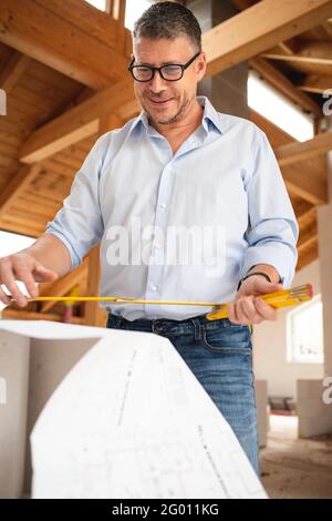 man with black glasses and blue shirt is looking at plan and is checking construction progress on building site in loft, attic in a house Stock Photo