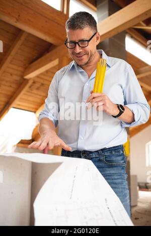 man with black glasses and blue shirt is looking at plan and is checking construction progress on building site in loft, attic in a house Stock Photo