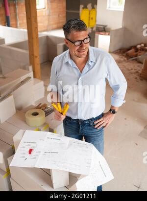 man with black glasses and blue shirt is looking at plan and is checking construction progress on building site in loft, attic in a house Stock Photo