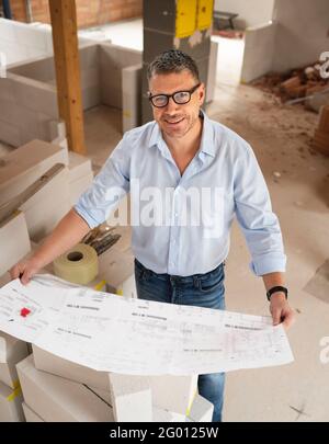 man with black glasses and blue shirt is looking at plan and is checking construction progress on building site in loft, attic in a house Stock Photo