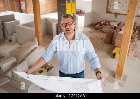 man with black glasses and blue shirt is looking at plan and is checking construction progress on building site in loft, attic in a house Stock Photo
