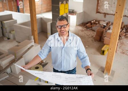 man with black glasses and blue shirt is looking at plan and is checking construction progress on building site in loft, attic in a house Stock Photo
