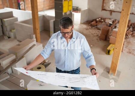 man with black glasses and blue shirt is looking at plan and is checking construction progress on building site in loft, attic in a house Stock Photo