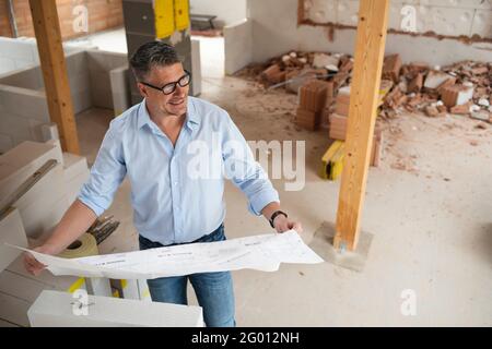 man with black glasses and blue shirt is looking at plan and is checking construction progress on building site in loft, attic in a house Stock Photo
