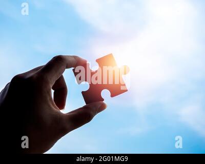 Jigsaw puzzle against blue sky background with copy space, silhouette. Hand of a businessman holding a piece of puzzle with sunlight. Business solutio Stock Photo