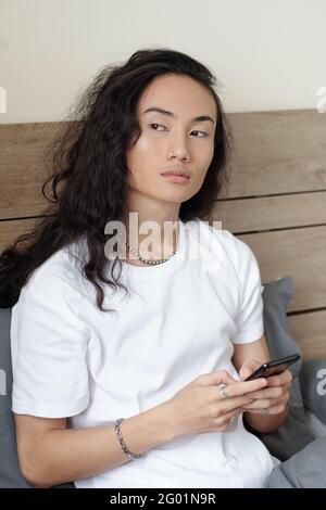 Serious long-haired Vietnamese guy with wavy black hair sitting on bed and looking away while texting message on gadget Stock Photo