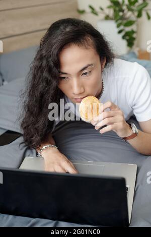 Serious young Asian freelancer with long hair lying on bed and eating bun while using laptop at home Stock Photo