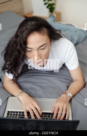 Serious young Vietnamese guy with wavy hair wearing earring in eye brow lying on bed and typing on laptop Stock Photo