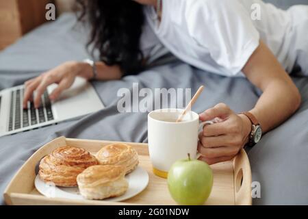 Close-up of unrecognizable man lying on bed and using laptop, he having breakfast in bed while working at home Stock Photo