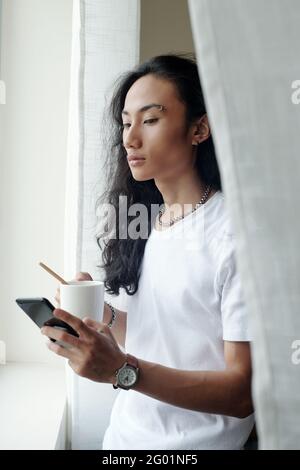 Serious young Vietnamese man with wavy hair standing at window and drinking coffee while scrolling through social media Stock Photo