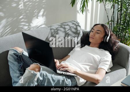 Serious modern young Asian guy in headphones lying on sofa and using laptop while working with social media at home Stock Photo
