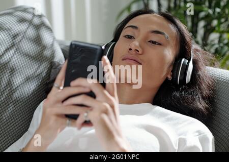 Serious handsome young Vietnamese man in wireless headphones lying on sofa and choosing music track on smartphone Stock Photo