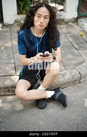 Serious handsome young Asian man with curly hair sitting on curb and listening to audio message in earphones outdoors Stock Photo