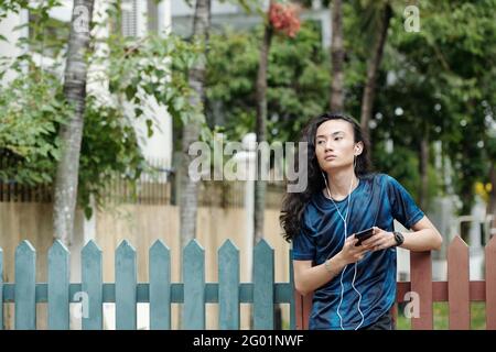 Serious handsome brunette young Asian man in earphones standing at fence and using smartphone while listening to music Stock Photo