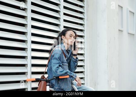 Serious handsome young Vietnamese man with long black hair sitting on bicycle and calling by phone while waiting for friend outdoors Stock Photo