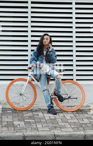 Serious pensive young Vietnamese guy with black long hair sitting on bicycle against shutter wall of building and talking by phone Stock Photo