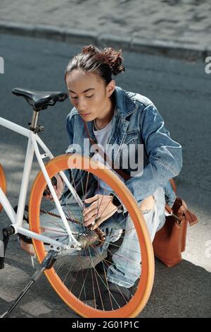 Serious young Vietnamese man with ponytail crouching on road and finding out reason of bicycle breakdown Stock Photo