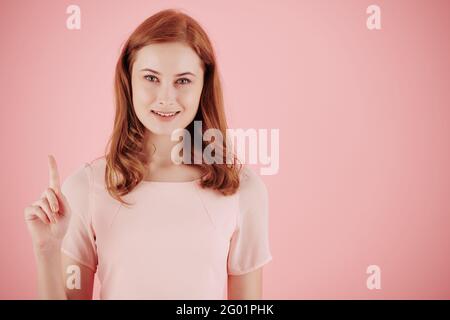 Beautiful young cheerful red haired woman pointing up with index finger and smiling at camera Stock Photo