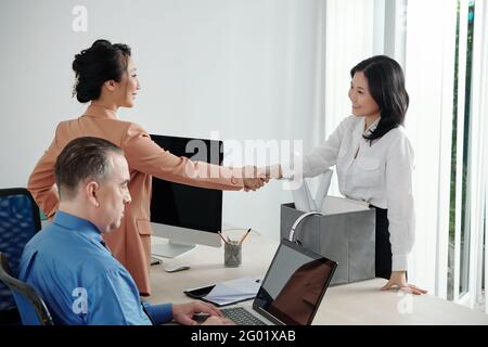 Cheerful pretty young businesswoman shaking hand of colleagues at her new workplace Stock Photo