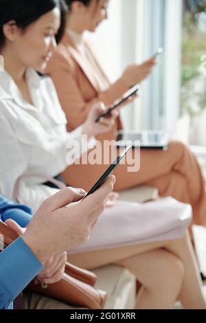 Business people sitting on chairs when waiting in queue and reading articles or checking social media on smartphones Stock Photo
