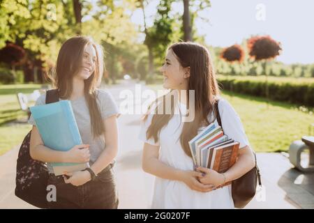 Two happy smiling student girls are walking and talking to each other on campus at sunset with warm light. Soft selective focus. Stock Photo
