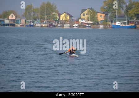 Broad-shouldered strong man is sailing in a single canoe on the bay. A professional athlete trains against the backdrop of various moored yachts. Prof Stock Photo