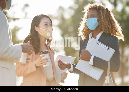 Positive entrepreneurs standing outdoors in medical masks and discussing details of project development during coffee break Stock Photo