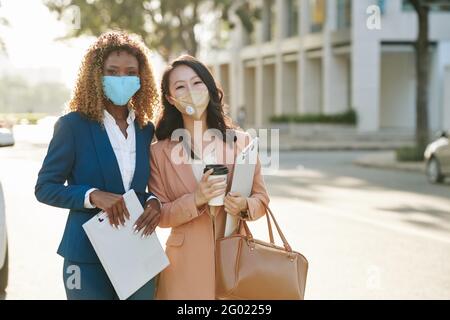 Pretty young cheerful businesswomen in protective masks standing outdoors with clipboards after important business meeting Stock Photo