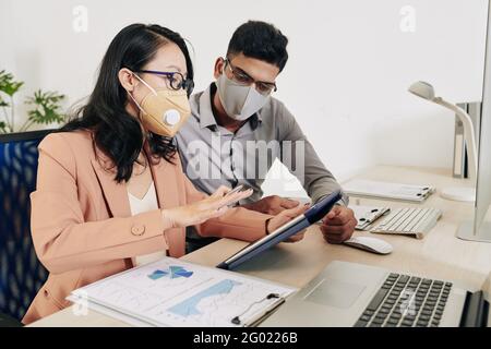 Serious businesswoman in protective mask and glasses showing presentation of her idea on tablet computer to colleague Stock Photo