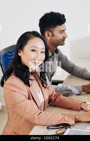 Pretty smiling young woman sitting at office desk next to her colleague and working on laptop Stock Photo