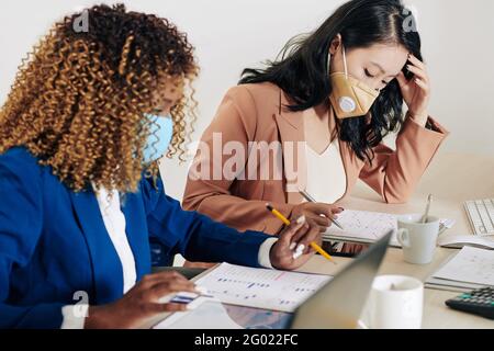 Serious concentrated businesswomen in protective masks analyzing financial reports when working with various charts at office desk Stock Photo