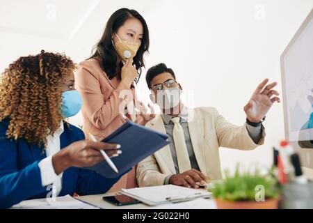 Business team in protective masks looking at chart on computer screen and discussing sales growth Stock Photo