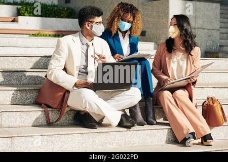 Multi-ethnic business team in protective masks sitting on steps outdoors and discussing charts and reports, planning work and project development Stock Photo