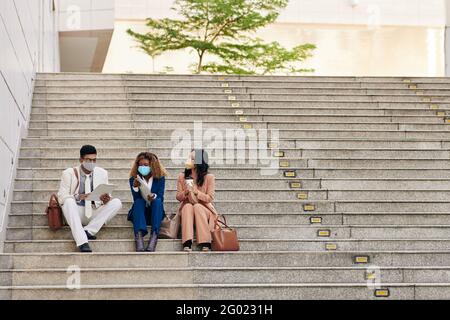 Group of young entrepreneurs in protective masks sitting on steps and discussing information in business documents Stock Photo