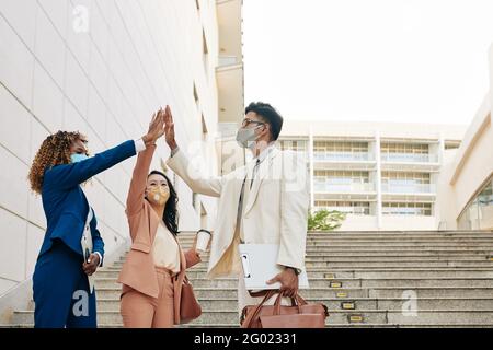 Group of happy excited business people in medical masks standing outdoors and giving each other high five after finishing work on big project Stock Photo