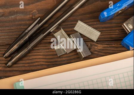 Ballpoint pen, stapler, and stapler staples a checkered notebook sheet on a wooden table. Close-up, selective focus. Self-development during self-isol Stock Photo