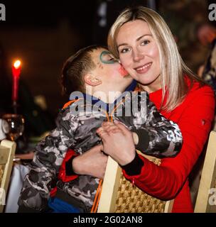 Happy Ukrainian mother with shining face and smile enjoys warm April evening with her son, who plays Indian with face painted mask, giving her a kiss Stock Photo