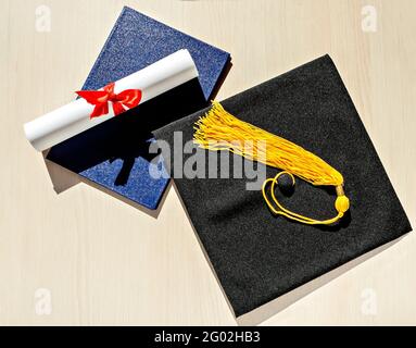 Black graduate cap with yellow tassel, blue diploma and paper scroll tied with red ribbon with bow on beige wooden background, Flat lay, top view, mor Stock Photo