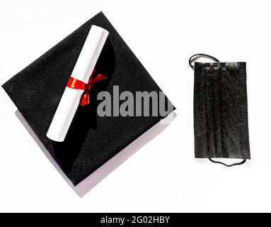 Black graduate cap paper scroll with red ribbon bow and protective face mask on white background, protection from virus, getting diploma in new realit Stock Photo