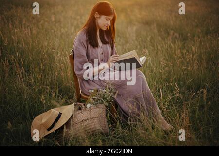 Beautiful woman in linen dress reading book in summer meadow in sunset. Young female with book and basket of flowers sitting on rustic chair and relax Stock Photo