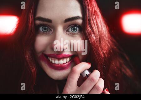 Portrait of crazy-looking teen girl with red hair she is smearing red lipstick on her face, horror concept. halloween time. Fear and nightmare Stock Photo