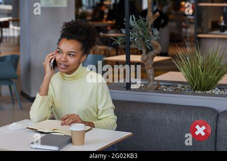 Portrait of young African-American woman speaking by smartphone while studying or working at cafe table with social distancing, copy space Stock Photo