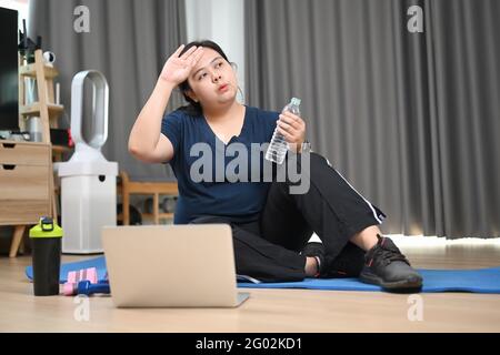 Tired young obese woman sitting and resting after doing fitness exercises at home. Stock Photo