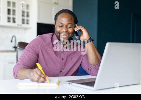 Cheerful and happy African-American male entrepreneur using laptop computer, talking on the smartphone and writes notes sitting at the desk at home, has phone conversation and makes schedule Stock Photo