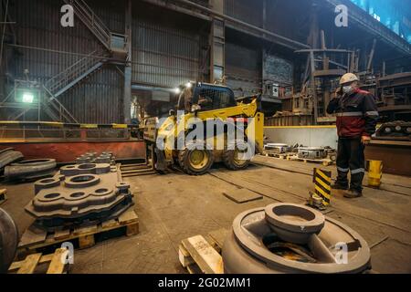 Workshop of metallurgical plant with small loading tractor or mower and unrecognizable engineer in protective mask, uniform and helmet. Stock Photo