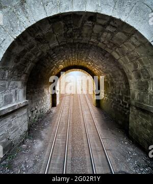 Dark passageway of roughly hewn stones with a round arch through which run two rail lines, composite surreal image Stock Photo