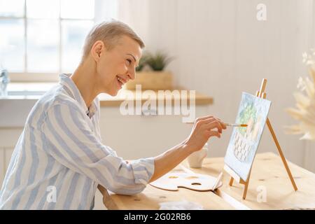 Creative hobby, digital detox. Side view of smiling mature woman painting picture at home while sitting at wooden table in kitchen, art therapy for mental health. Senior people and art concept Stock Photo