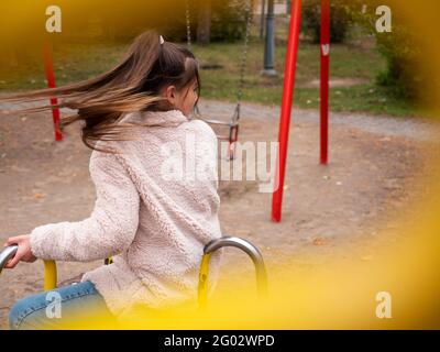 View through railings over a happy smiling teenage girl (11-12) in a faux fur coat and with long ponytail spinning at a merry-go-round at playground. Stock Photo