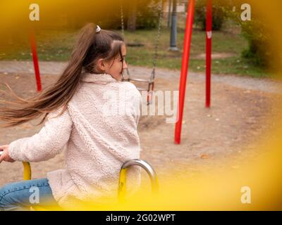 View through railings over a happy smiling teenage girl (11-12) in a faux fur coat and with long ponytail spinning at a merry-go-round at playground. Stock Photo